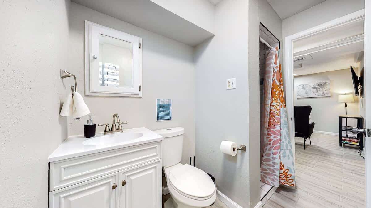 Un baño con lavamanos, espejo y retrete blancos. Hay una cortina de ducha con estampado floral.