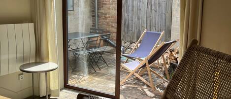 Une pièce comprenant un fauteuil en osier, une petite table et une porte-fenêtre coulissante donnant sur un patio extérieur avec une table et des chaises.