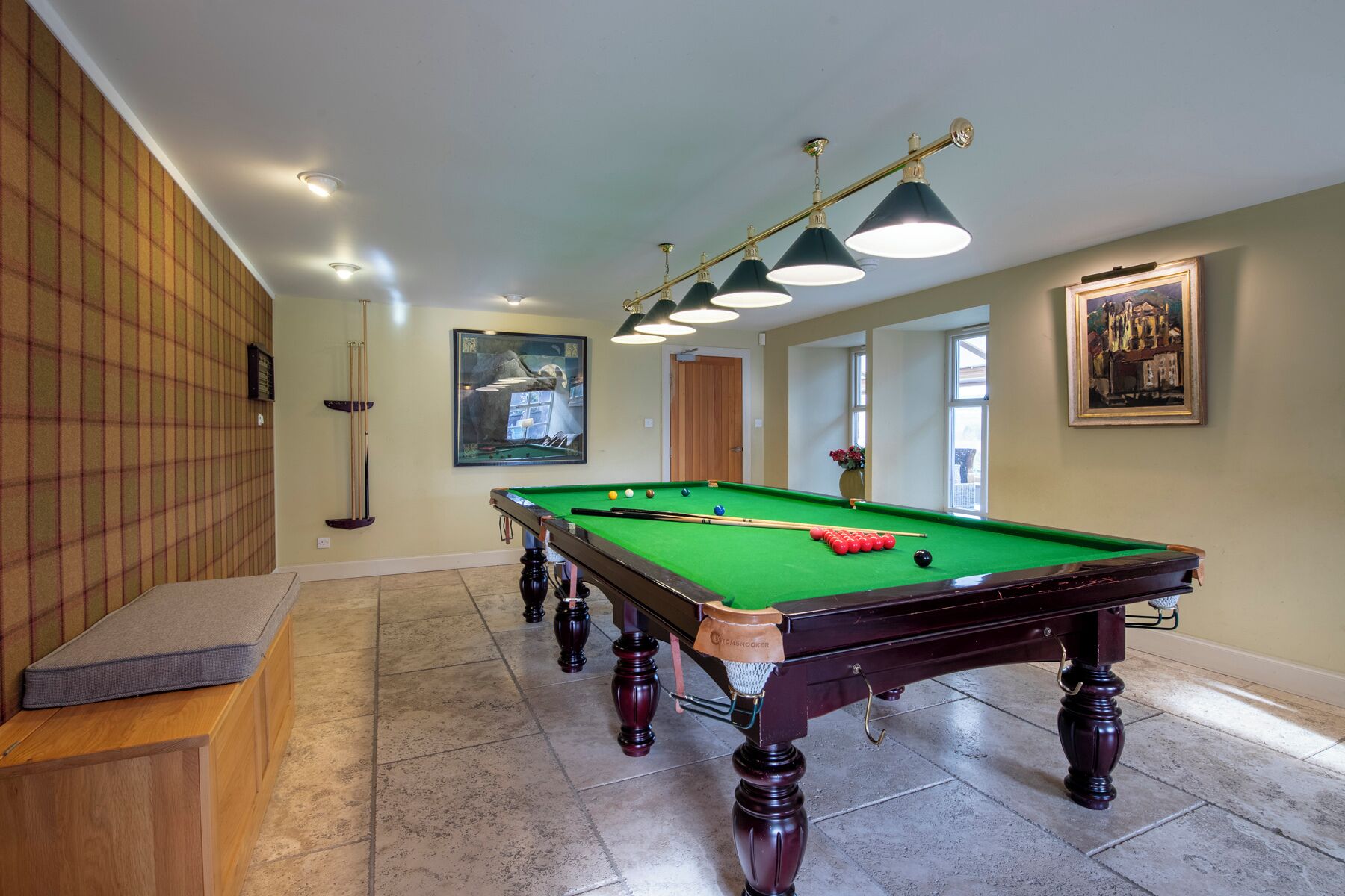A pool table with a green felt surface, surrounded by a wooden bench, a wall-mounted pool cue rack, and a ceiling light fixture.