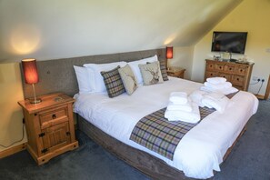 A neatly made bed with white linens and plaid blankets, a television on a wooden dresser, and a lamp on a bedside table.