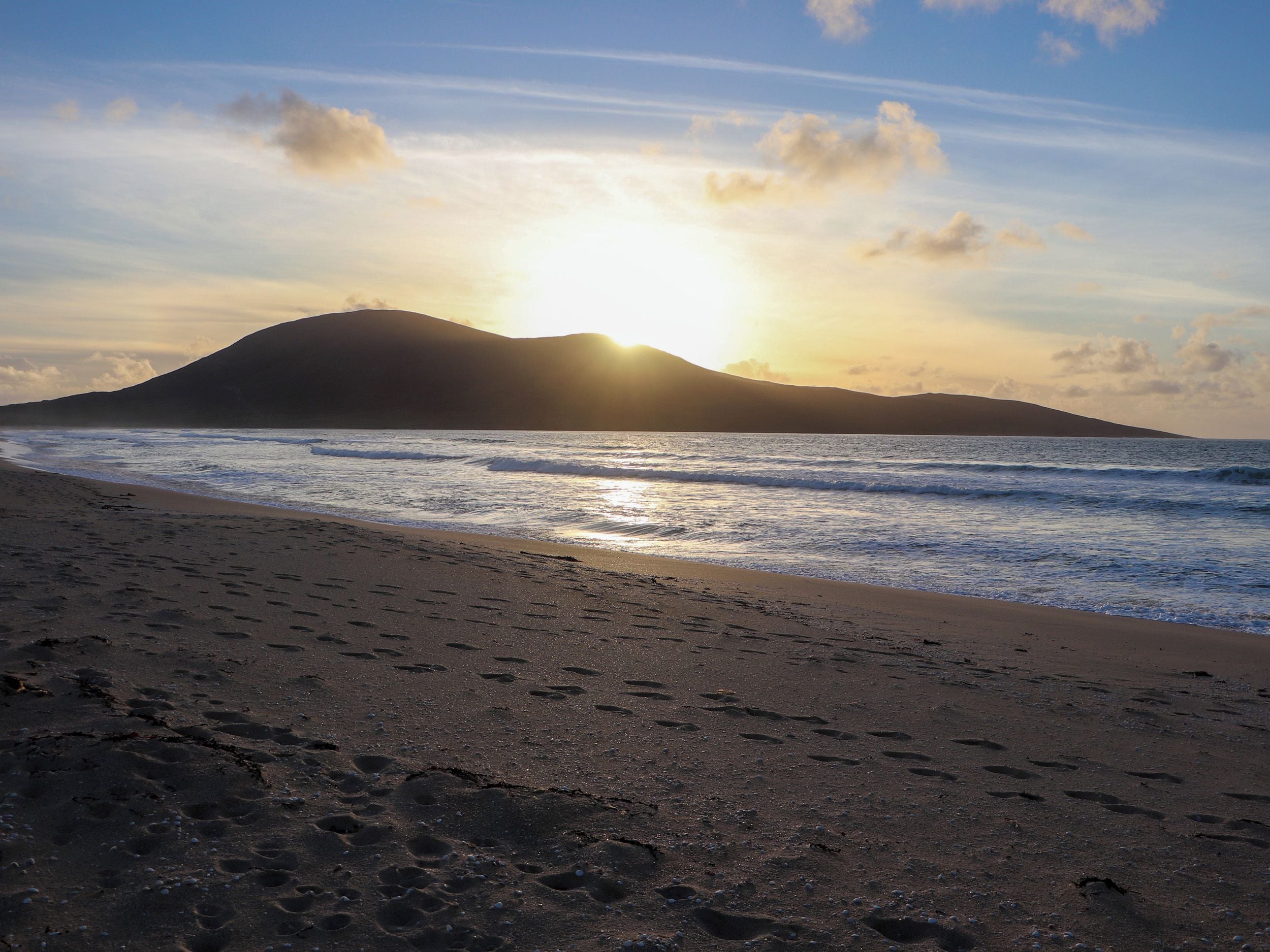A beach with footprints leading to the water, a mountain in the background, and the sun setting over the horizon.