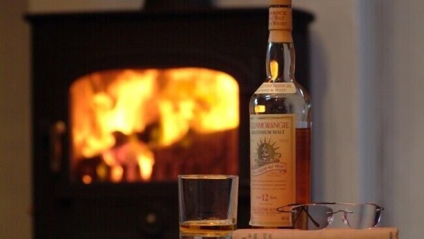 Een fles whisky, een shotglas en een paar brillen op een tafel, met een open haard op de achtergrond.