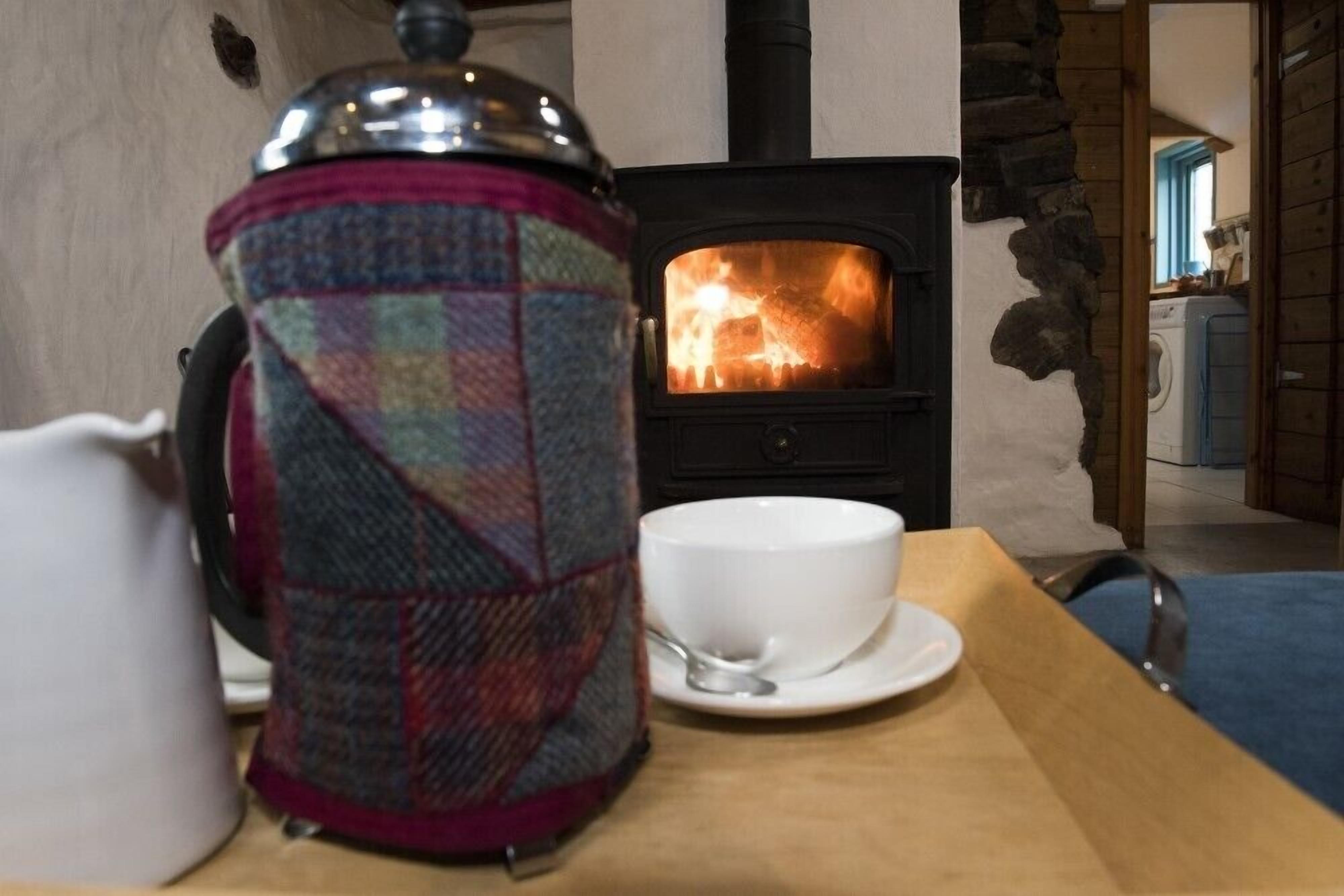 Una habitación acogedora con una estufa de leña, una tetera de estampado escocés y una taza blanca sobre una mesa de madera.