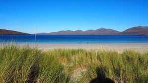 Een strand met helderblauw water, begroeide duinen en verre bergen.