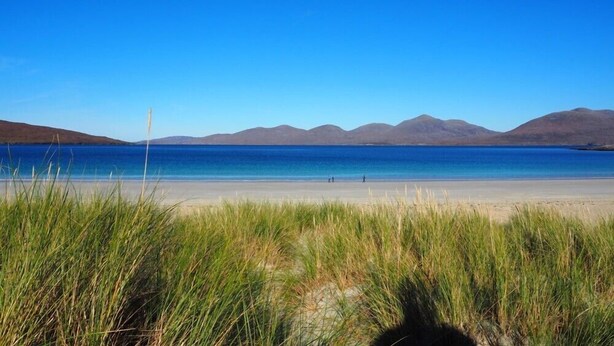 Una playa con aguas azules cristalinas, dunas cubiertas de hierba y montañas a lo lejos.