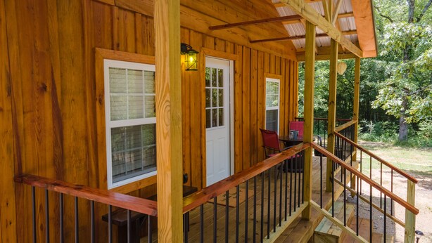 A wooden cabin with a porch, a dining area with a table and chairs, and a view of the surrounding forest. - Escape to cozy 2-bedroom cabin near Ouachita Forest 4 min drive away (Mena)