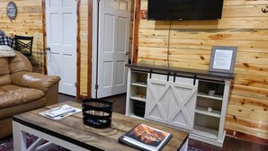 A cozy room with a leather sofa, a wooden coffee table with magazines, a TV mounted on the wall, and a white cabinet with shelves. - Escape to cozy 2-bedroom cabin near Ouachita Forest 4 min drive away (Mena)