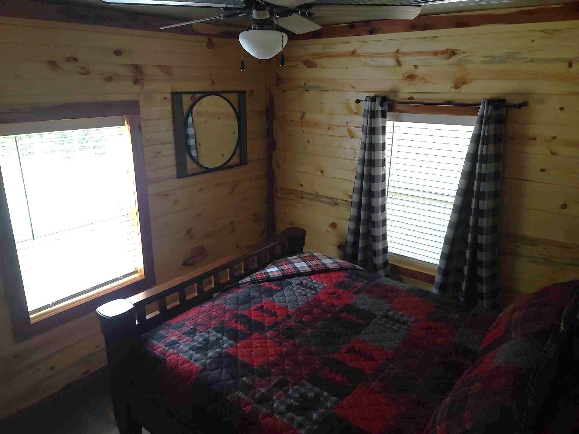Una habitación con cama, ventilador de techo, ventana con persianas y espejo adosado a la pared.