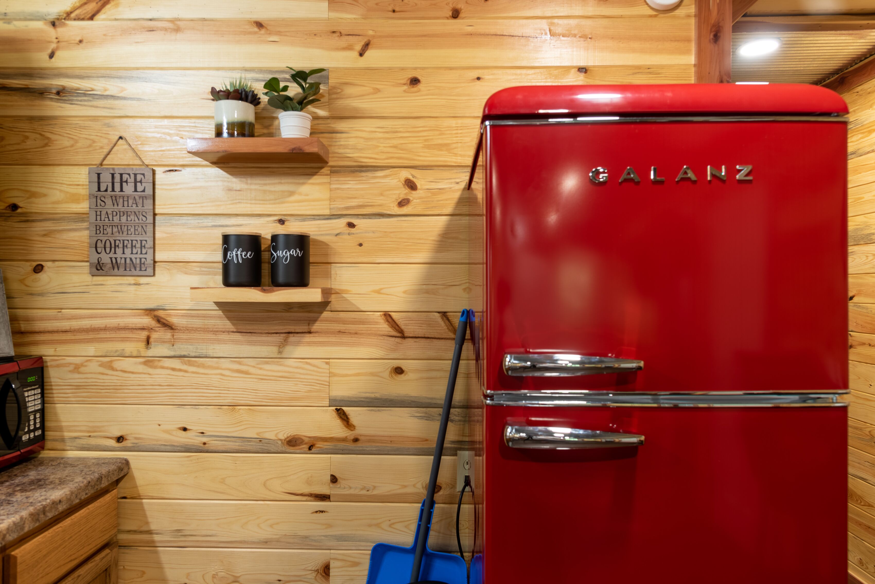 Una cocina con un refrigerador Galanz rojo, paneles de madera en la pared, un estante con plantas y tazas, y un letrero que dice 'Life is what happens between coffee & wine'.