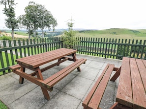 Ett picknickbord i trä står på en betongterrass, med ett grönt metallstaket och utsikt över böljande kullar och vindkraftverk i fjärran.