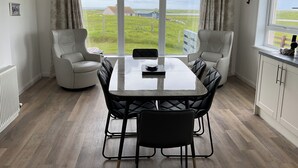A dining area with a table and chairs, large windows with curtains, and a view of a grassy landscape. - Fuidaigh (South Uist)