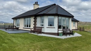 A house with a pitched roof, large windows, and a patio area with benches and a table. - Fuidaigh (South Uist)