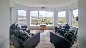 A spacious living room with a large bay window offering a scenic view of a lake and hills. There are two dark armchairs with patterned cushions and a small wooden table with a vase of flowers. - Fuidaigh (South Uist)