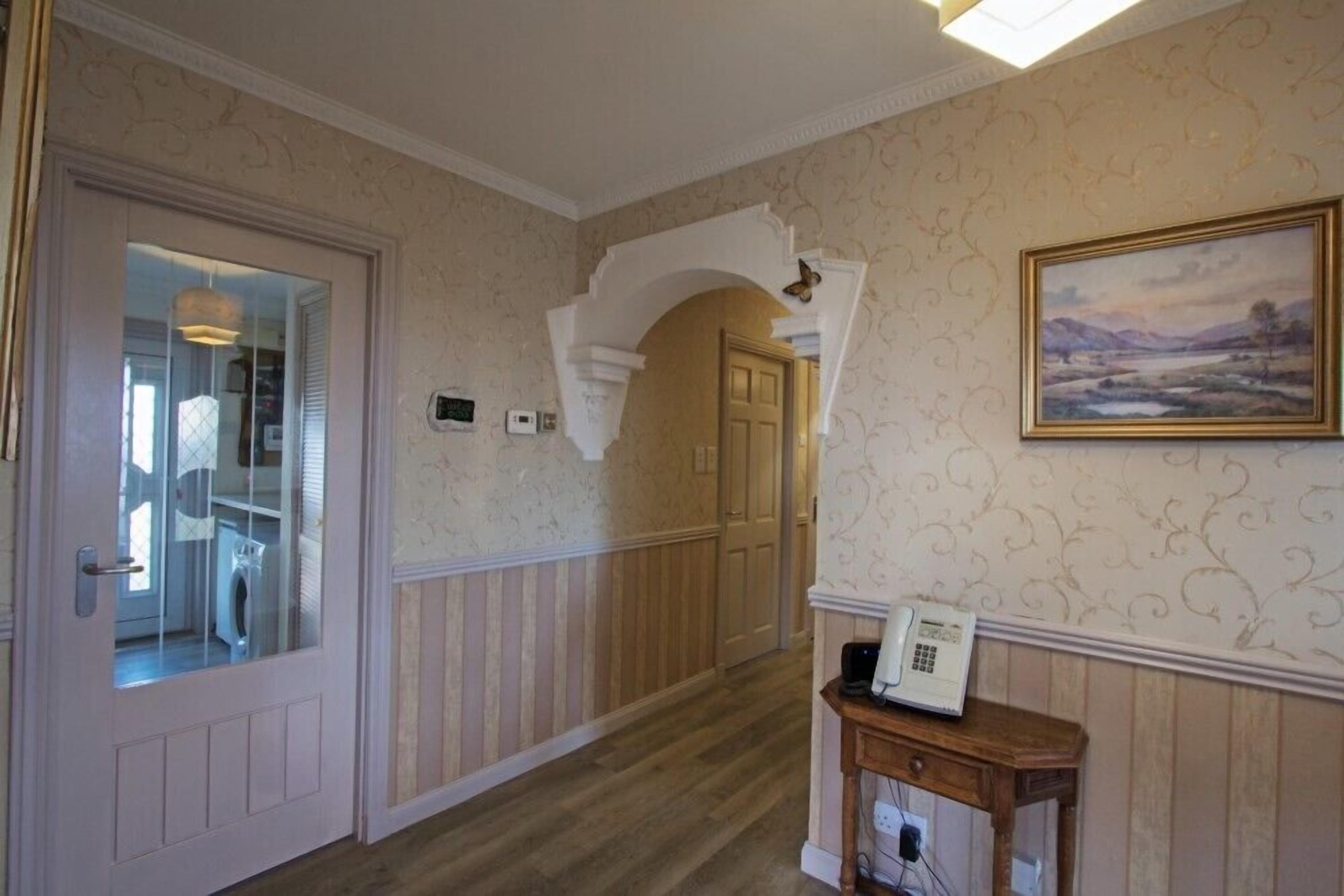 A hallway with a wooden floor, a white door with a glass panel, a telephone on a wooden table, and a painting on the wall.