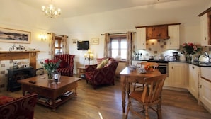 A spacious living room with a wooden coffee table, red armchairs, and a dining table with chairs. A kitchen with wooden cabinets and a stove is visible in the background. - Smiddy Cottage (South Uist)