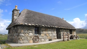 A stone house with a thatched roof, a wooden wheel, and a stone chimney. - Smiddy Cottage (South Uist)