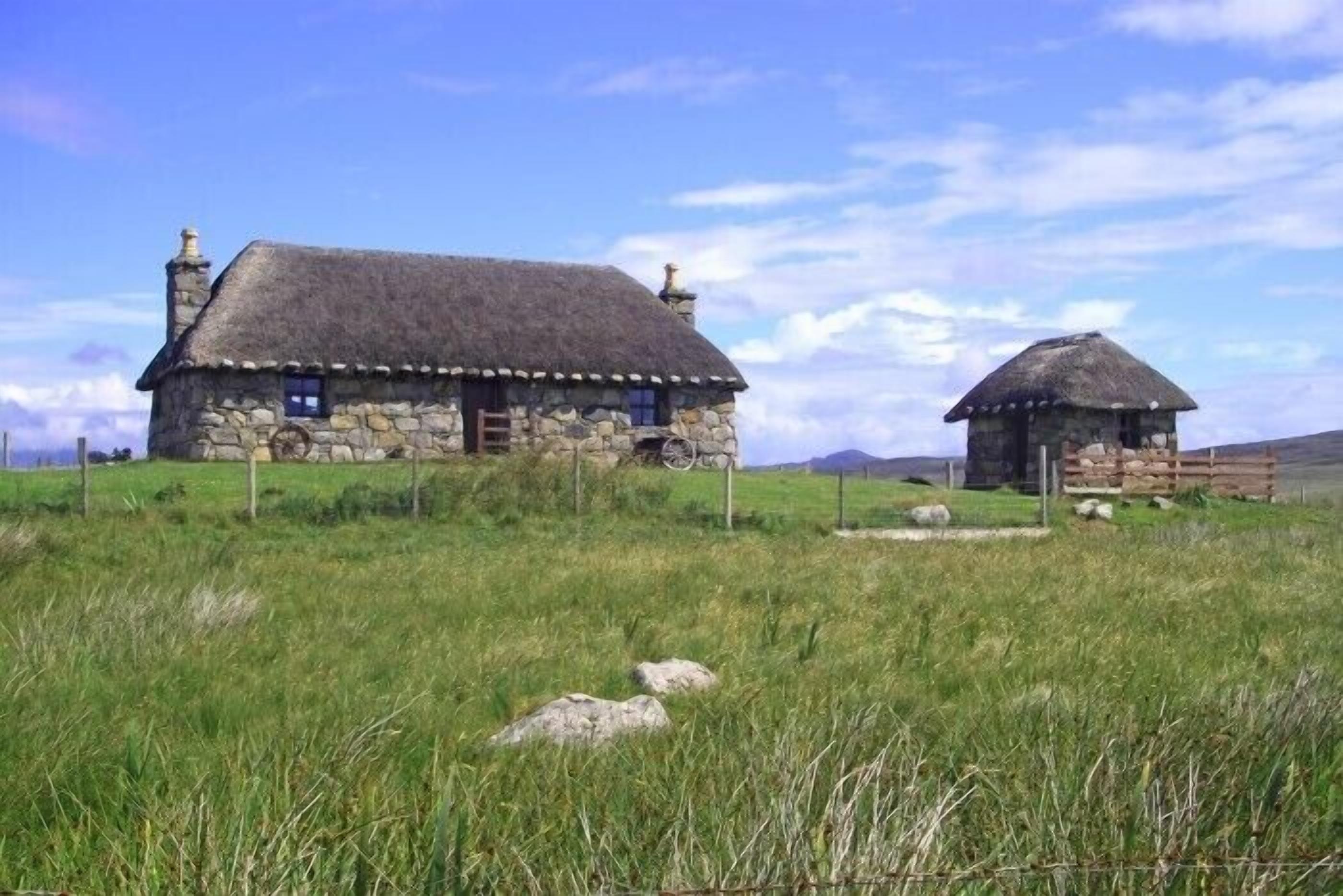 Une maison en pierre avec un toit de chaume, entourée d’un champ verdoyant et d’une petite construction en pierre.