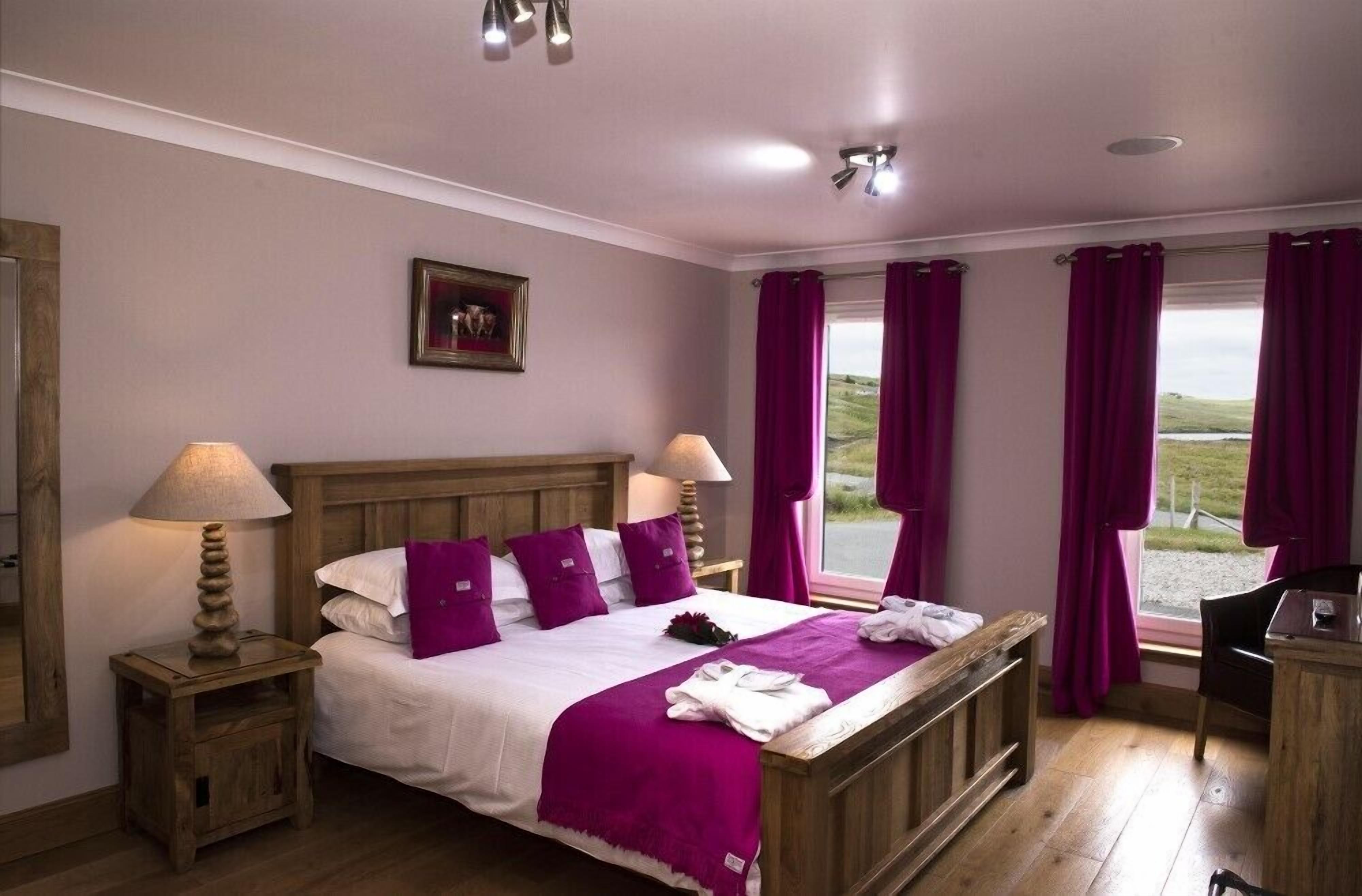 A bedroom with a wooden bed, two bedside tables with lamps, a view of the outdoors, and a framed picture on the wall.