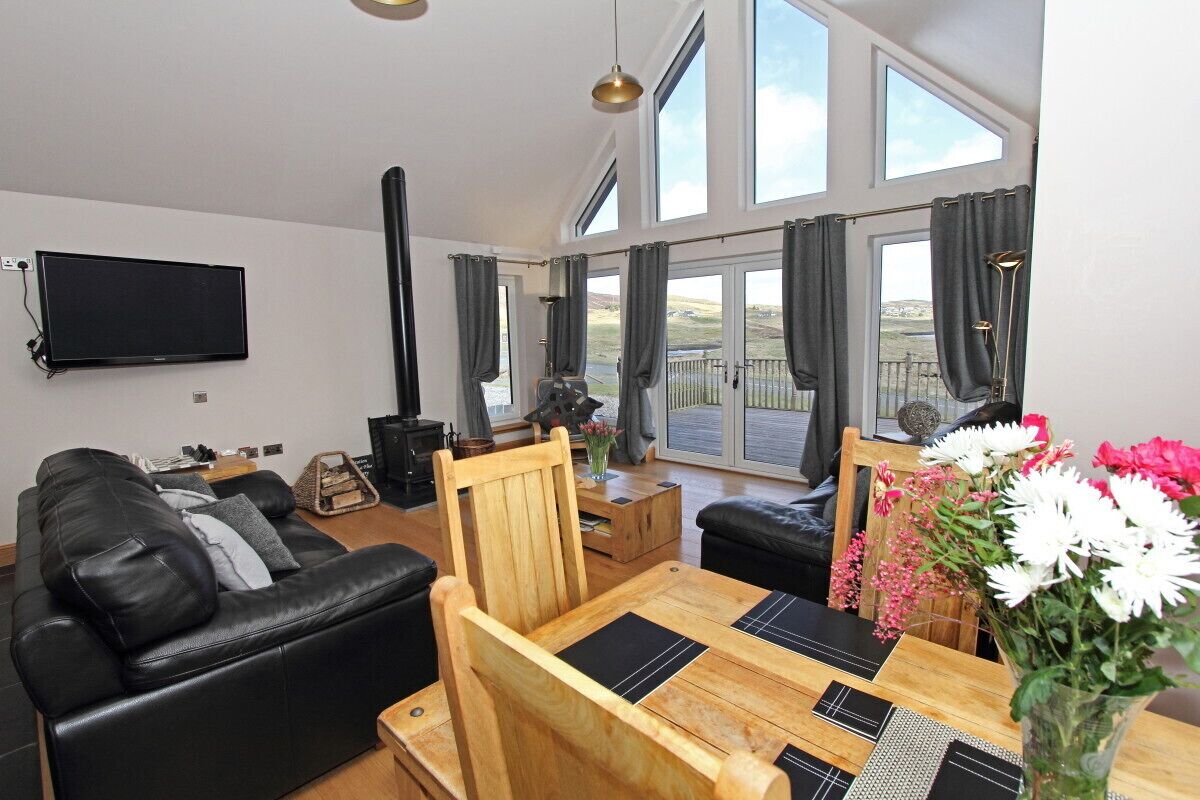 A modern living room with a black leather sofa, a wooden dining table, and a large window with curtains.