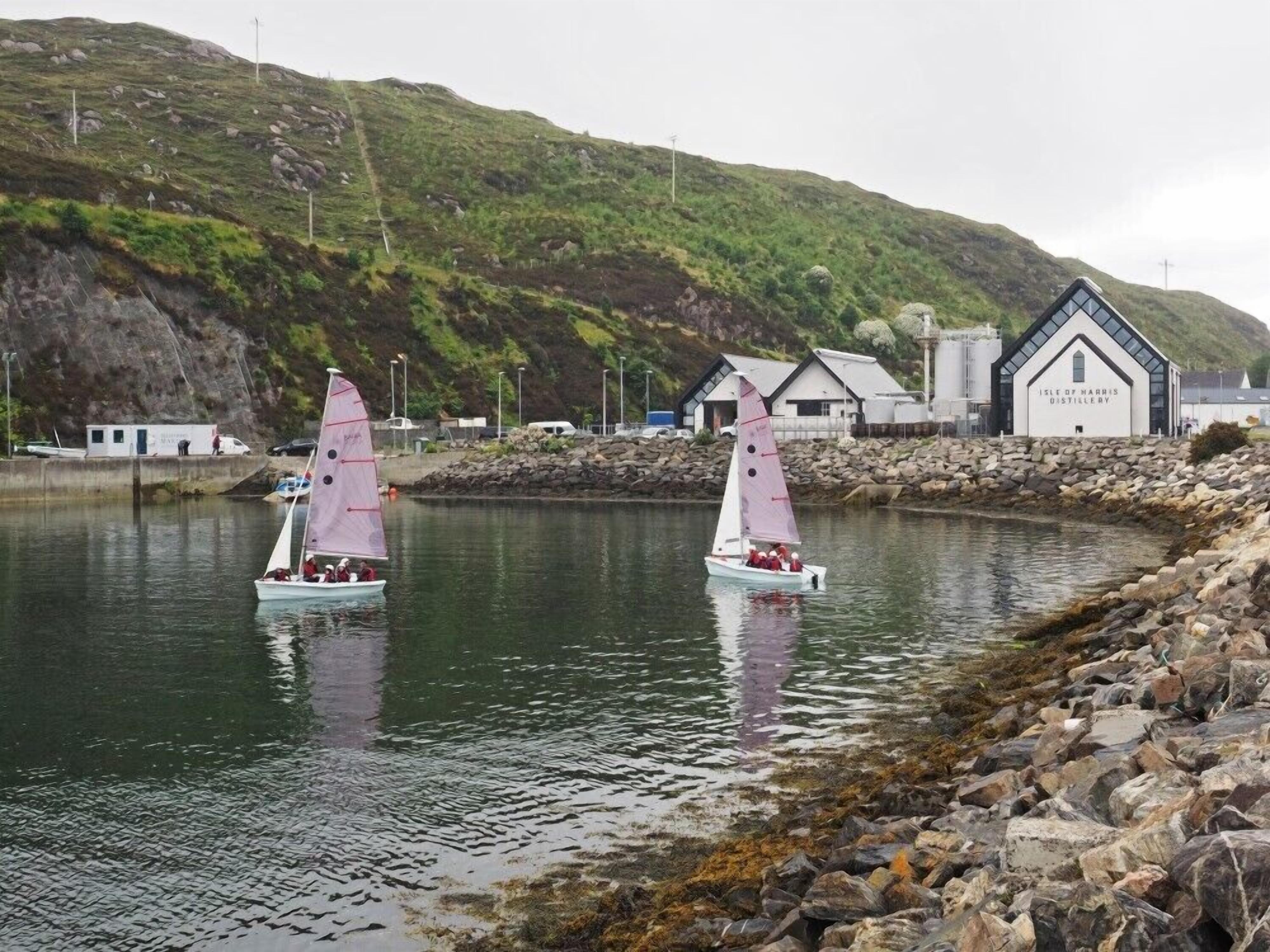 Dos veleros sobre aguas tranquilas cerca de una costa rocosa con un edificio que dice 'Isle of Narnia Distillery'.