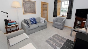 Una sala de estar con un sofá gris, un sillón gris, una mesita con una lámpara, un televisor y una estantería.