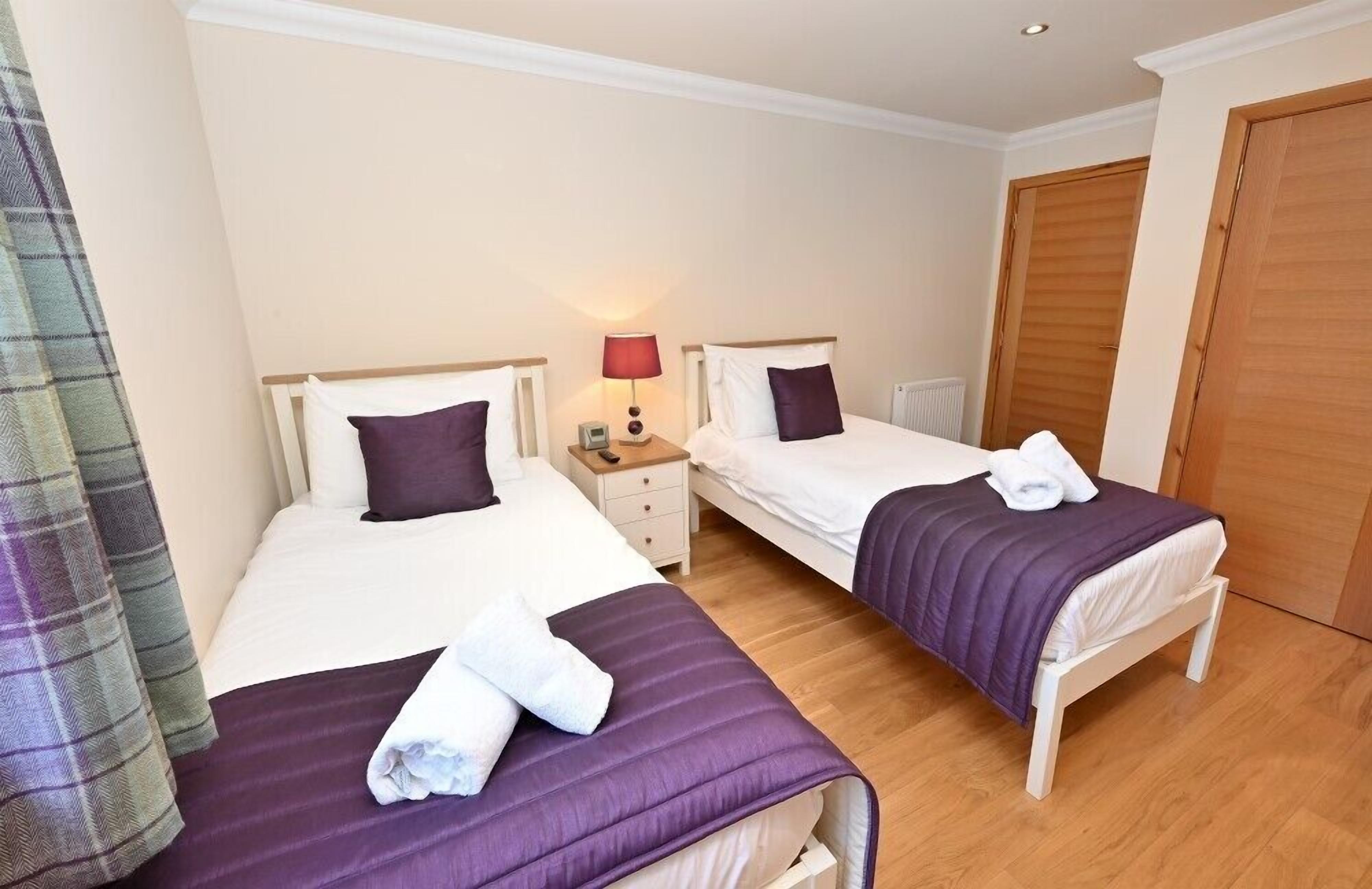 A room with two beds, each with a purple bedspread and white pillows. There is a nightstand with a lamp between the beds.