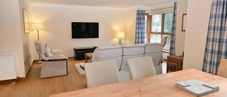 A spacious living room with a wooden dining table, a grey sofa, and a flat-screen TV mounted on the wall.
