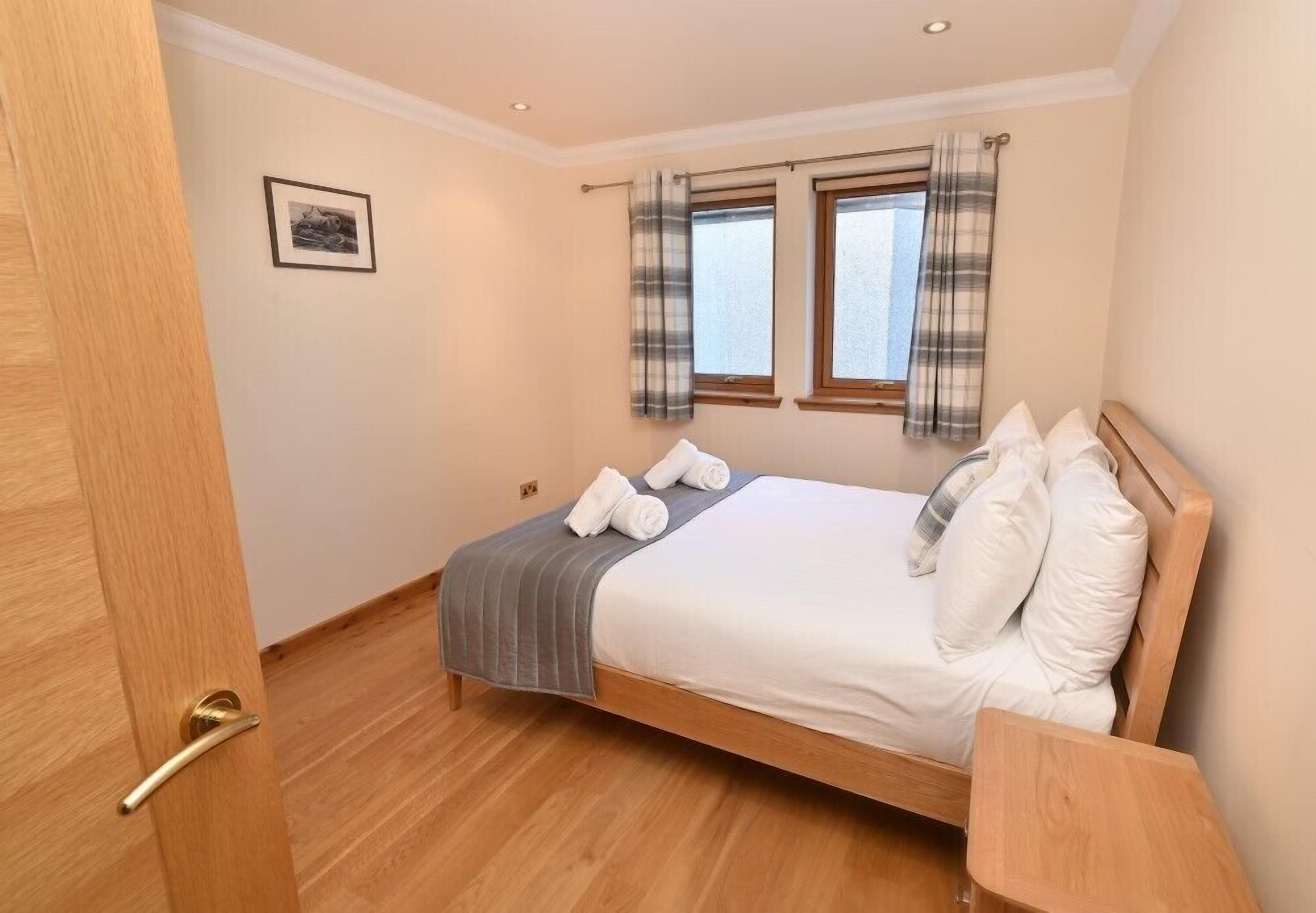 Ein Schlafzimmer mit einem hölzernen Bett, einem Nachttisch und zwei mit Vorhängen versehenen Fenstern.