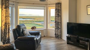 A living room with a leather sofa, a TV, and large windows with curtains. - Lingeigh (South Uist)