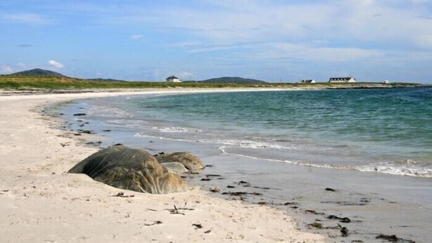 A sandy beach with clear water, a large rock in the foreground, and a few buildings in the distance. - Lingeigh (South Uist)