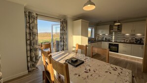 A dining area with a table and chairs, a kitchen with modern appliances, and a view of the countryside through a glass door. - Lingeigh (South Uist)