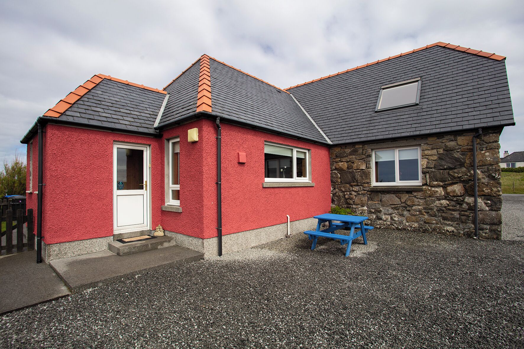 A two-story building with a red exterior and stone accents, featuring a gravel driveway and a blue picnic table.