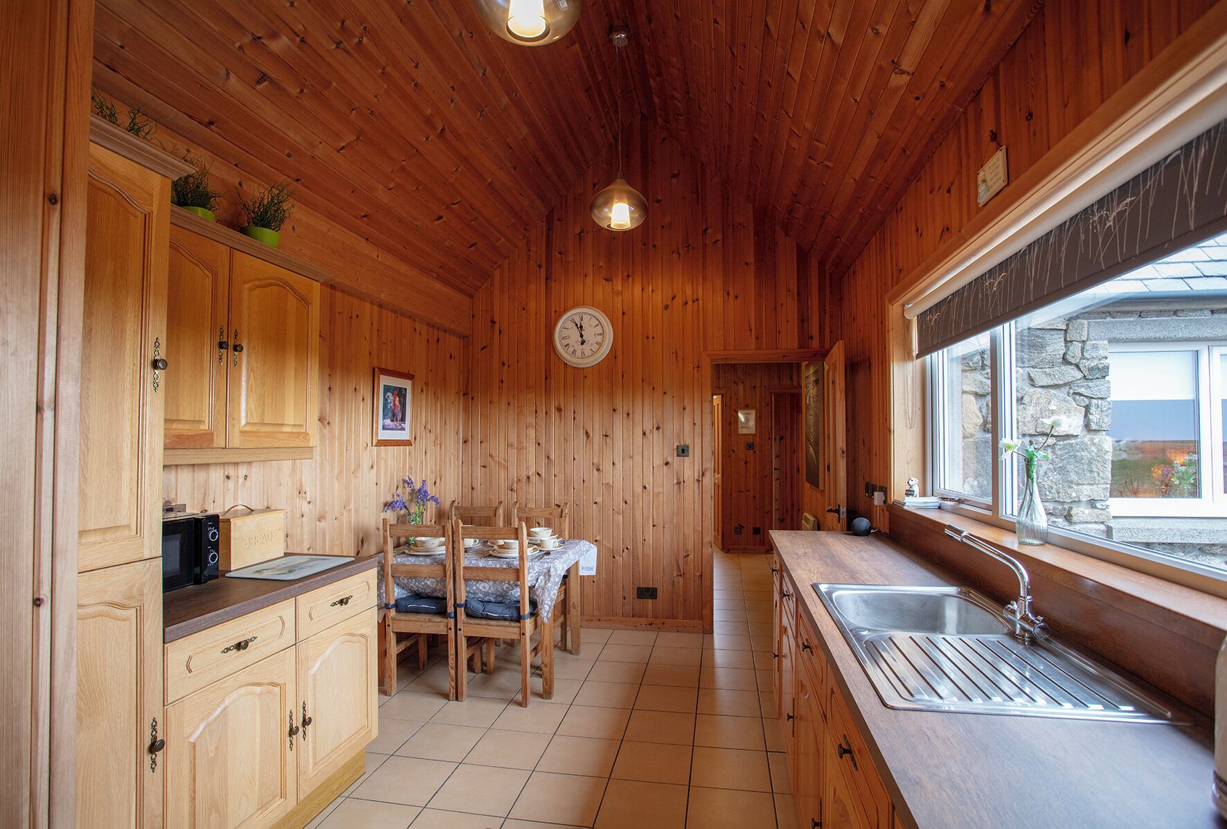 A wooden interior with a kitchen area, including cabinets, a dining table with chairs, a clock, and a window with a view.
