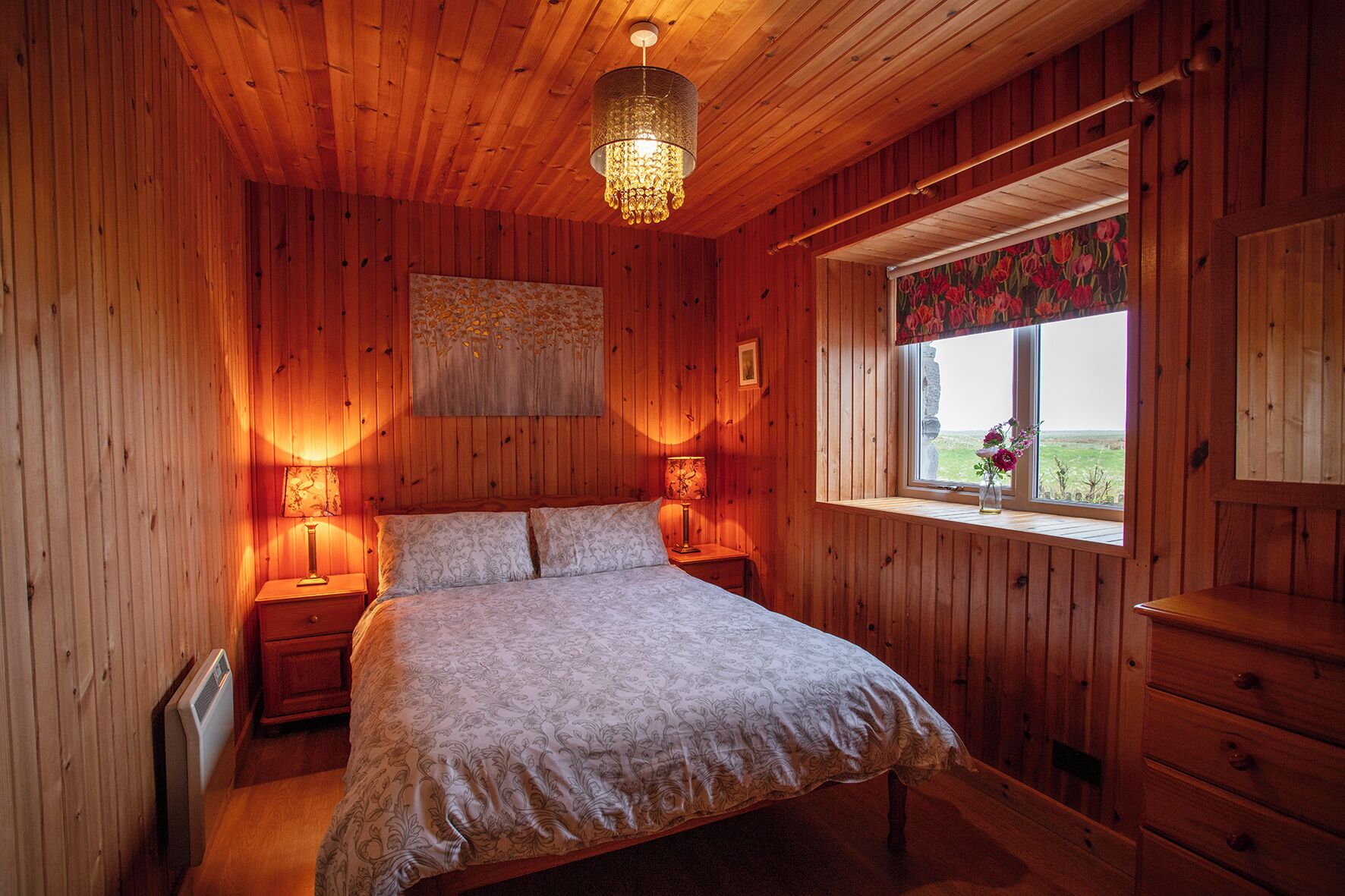 Une chambre à coucher aux murs lambrissés, avec un lit, deux lampes de chevet, un lustre, une fenêtre donnant sur un jardin fleuri et un radiateur.