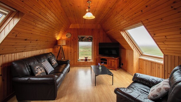A cozy attic room with wooden walls and sloped ceilings, featuring a leather sofa, a small coffee table, and a television. - Kelp Cottage (South Uist)