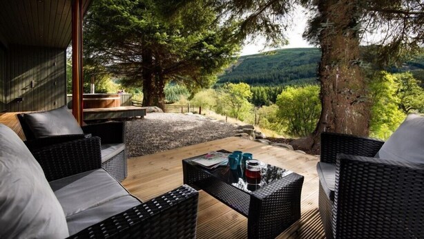 Terraza de madera con mobiliario de mimbre y una mesa de vidrio, con vistas a un valle boscoso.