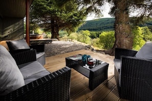 Terraza de madera con mobiliario de mimbre y una mesa de vidrio, con vistas a un valle boscoso.