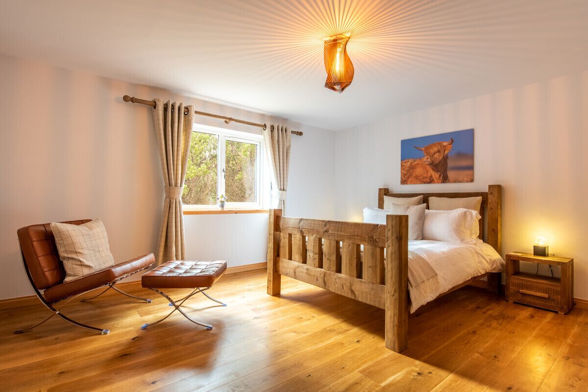 Une pièce avec un lit en bois, un canapé en cuir, une table basse en bois, une fenêtre avec des rideaux et une lampe fixée au mur.