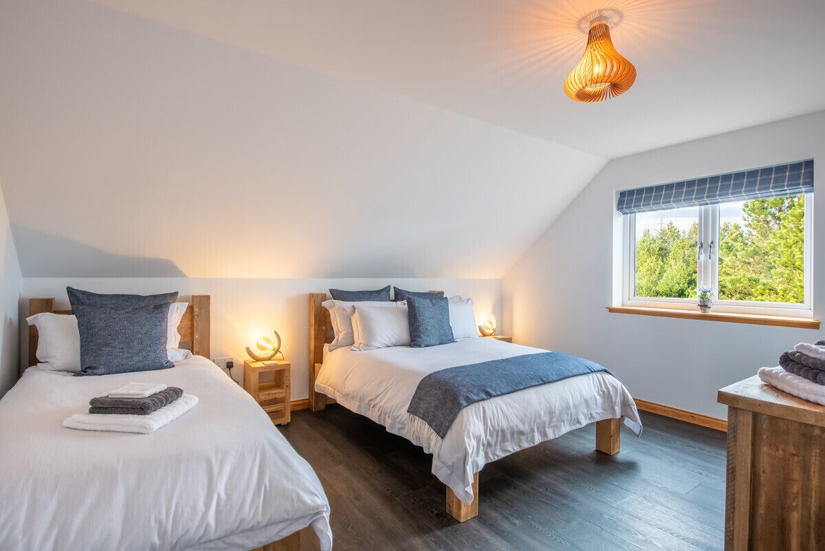 Quarto com duas camas, cabeceiras de madeira, mesas de cabeceira com luminárias, janela com vista para áreas verdes e luminária no teto.