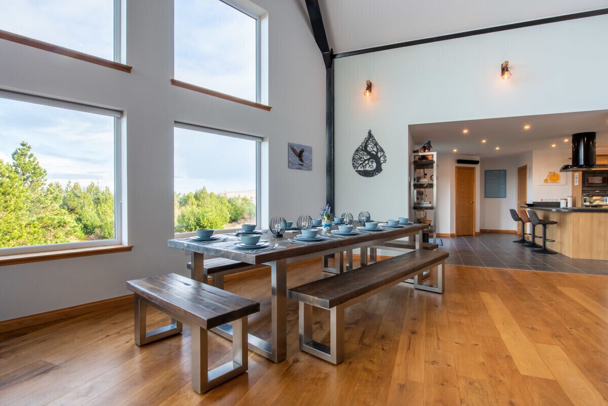 Un comedor amplio con una larga mesa de madera dispuesta para una comida, grandes ventanas que ofrecen vistas a la vegetación, y una cocina moderna al fondo.