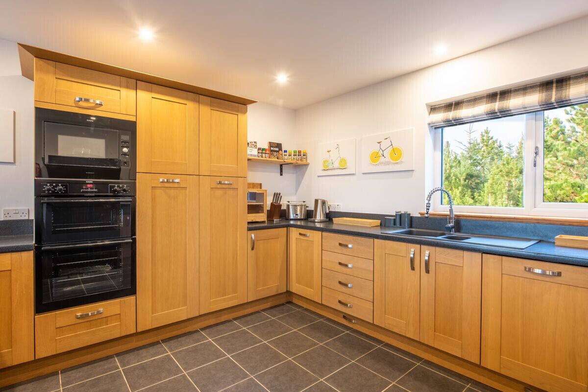 A modern kitchen with wooden cabinets, a built-in oven, a black countertop, and a window with a view of trees.