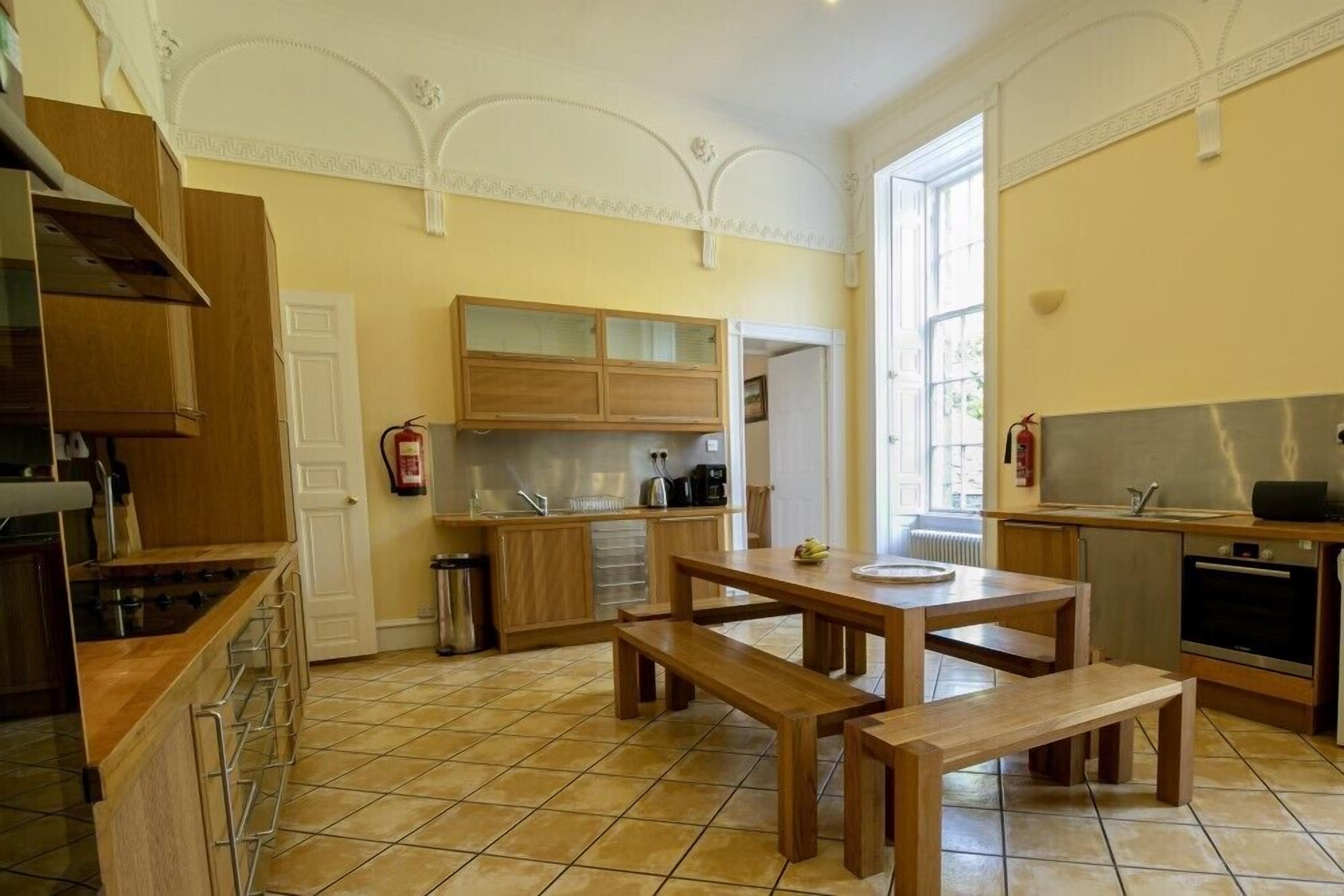 Une cuisine avec des meubles en bois, une table à manger avec des bancs, une cuisinière et une fenêtre laissant entrer la lumière naturelle.