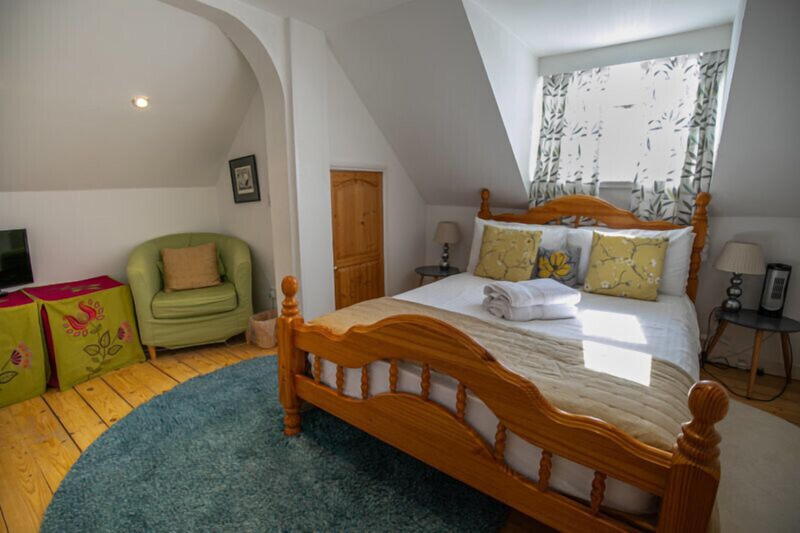 Un dormitorio con una cama de madera, un sillón verde, un armario de madera y una ventana con cortinas estampadas.