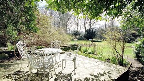 An outdoor patio with a round table and white chairs, surrounded by lush greenery and trees. - Cortes House (Fraserburgh)