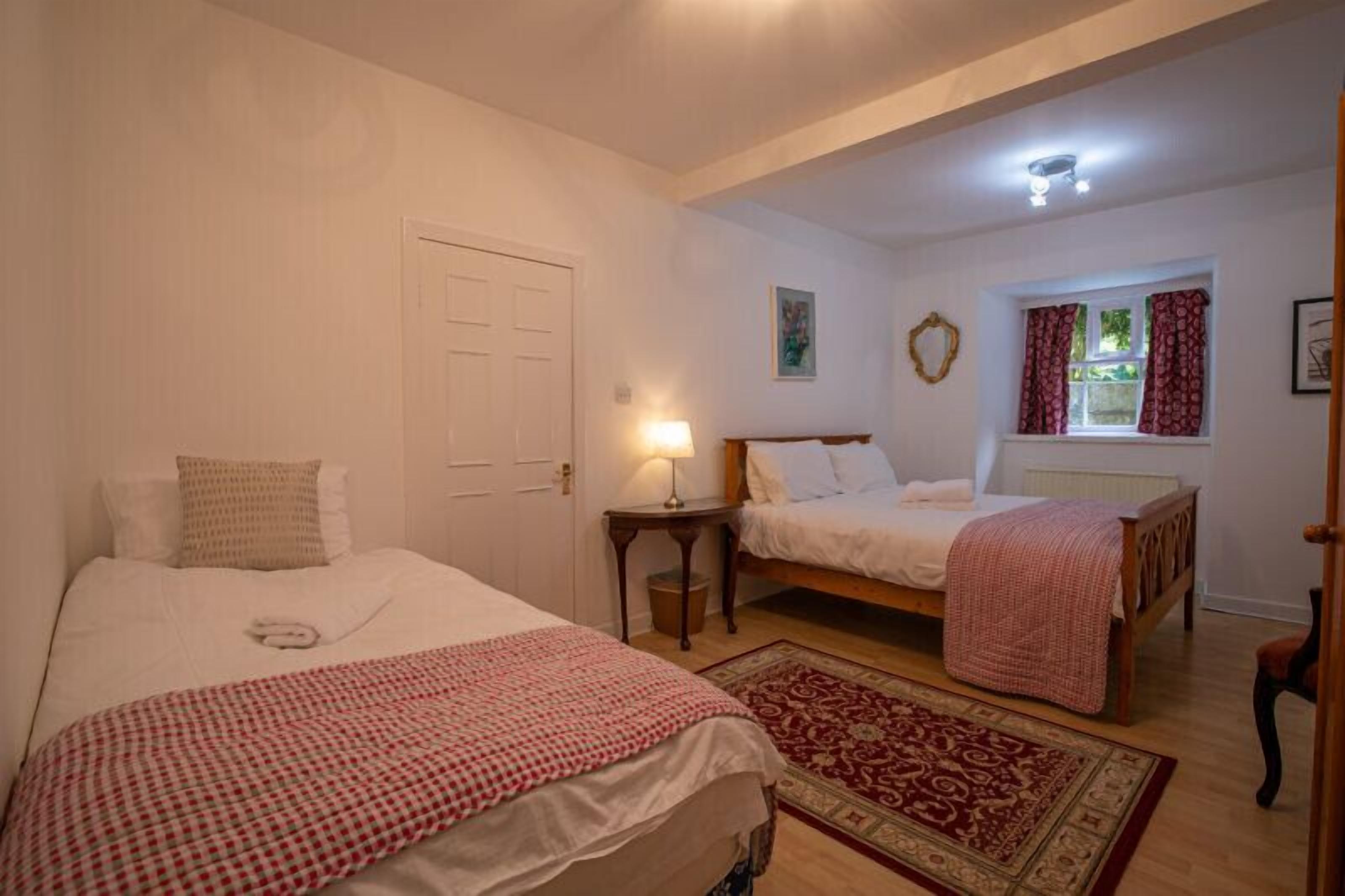 A bedroom with two single beds, a bedside table with a lamp, a window with curtains, and a door.