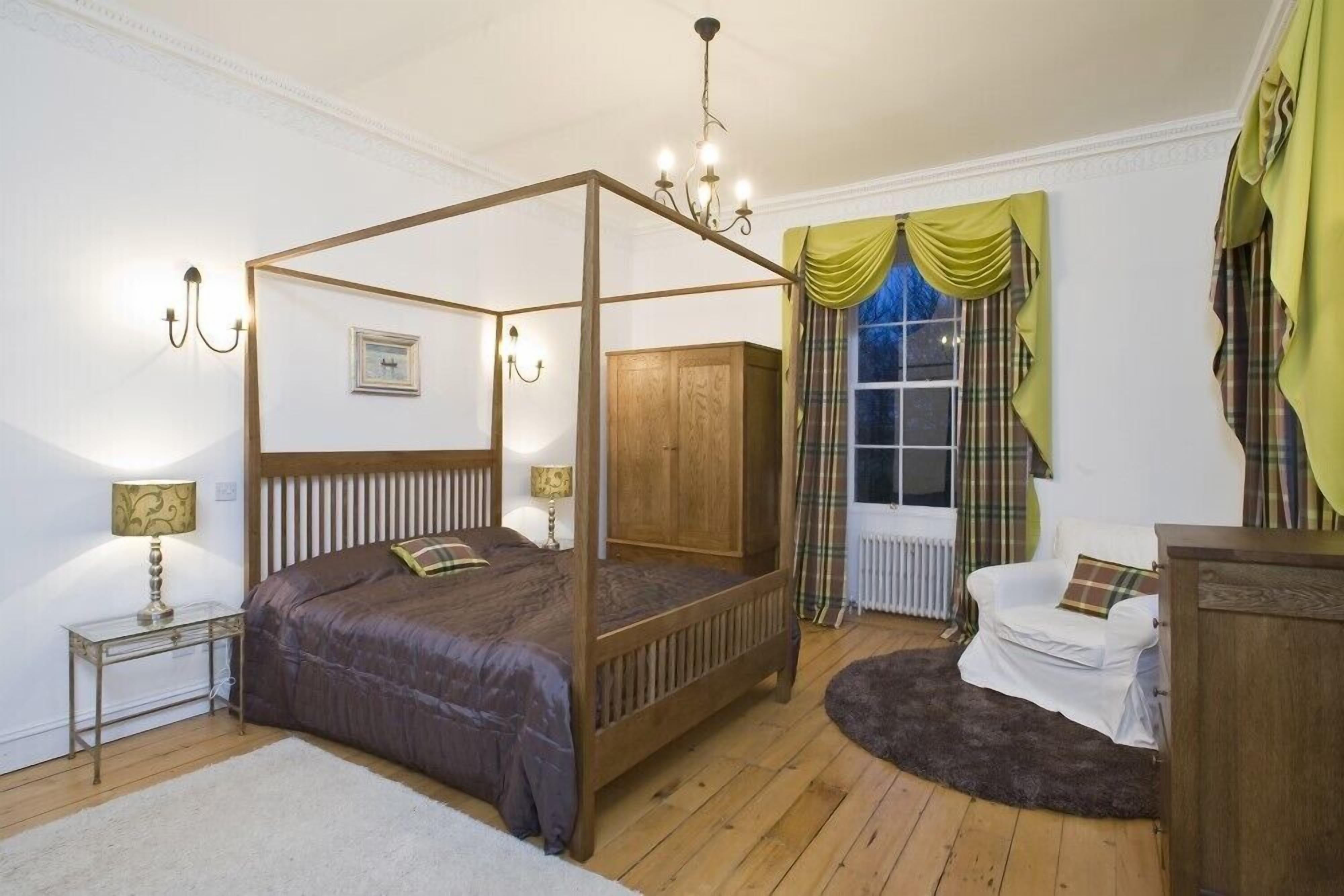 A four-poster bed with a wooden frame, a white radiator, a white armchair, and a small table with a lamp.