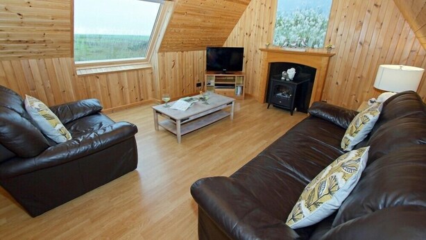 A cozy wooden cabin interior with a large window, a fireplace, a television, and leather sofas. - Canach Cottage (South Uist)