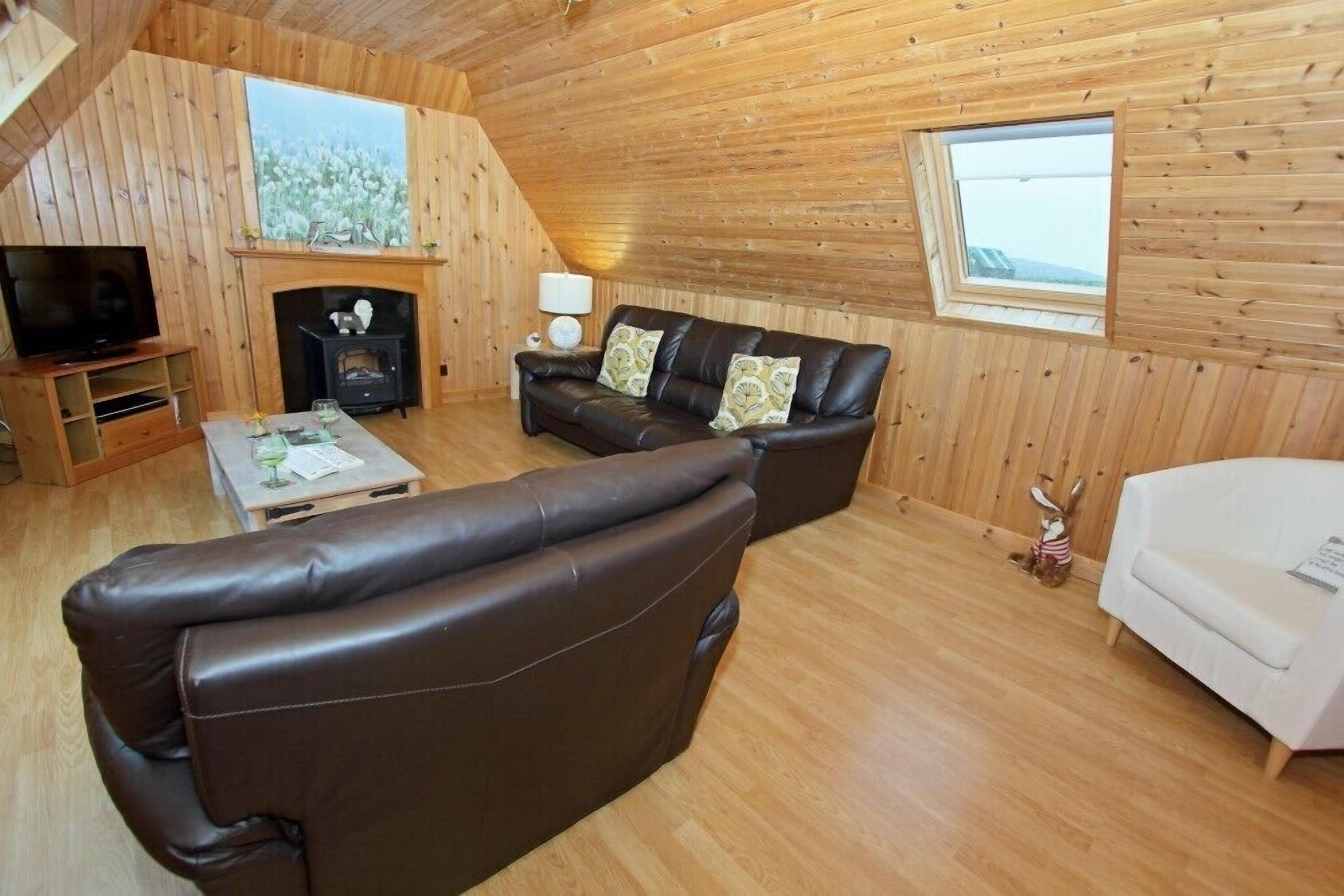 A cozy cabin interior with a fireplace, leather sofas, a television, and a window overlooking a snowy landscape.