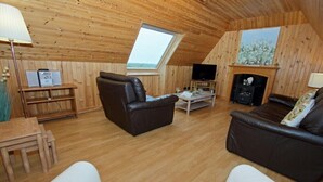 A cozy loft-style room with a wooden ceiling, a fireplace, a television, and two leather sofas. - Canach Cottage (South Uist)