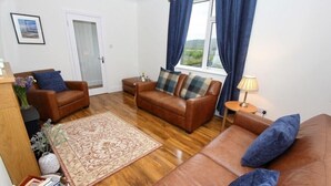 A living room with brown leather sofas, a patterned rug, a wooden floor, a door, and a window with curtains. - Tulloch House (South Uist)